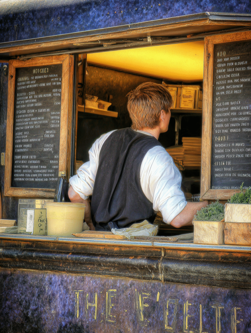 A young man working in a food truck has his back to the camera as he places an order to the kitchen through the food truck window.